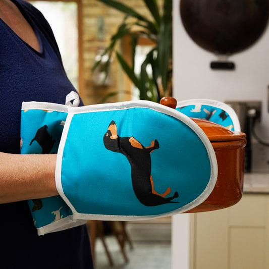 Person carrying a casserole dish using a blue oven mitt with a dachshund design in a kitchen.