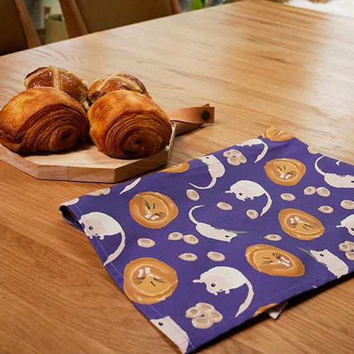 Baked pastries on a wooden board with a purple tea towel featuring dormouse design on a wooden table.