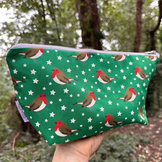 A green zippered pouch with a white star pattern and red-breasted robin birds, held in a hand against a blurred forest background.