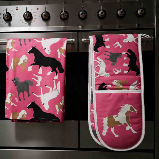 Pink oven gloves and tea towel with horse pattern shown hanging over the handle of a staninless steel stove.