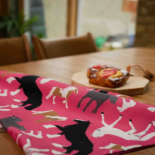 Pink tea towels with horse patterns on a wooden table with pastries in the background