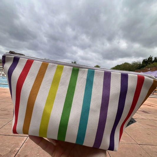 A rectangular zippered pouch with vertical rainbow stripes in red, orange, yellow, green, teal, and purple on a white background, held in a hand in front of a lido outdoor swimming pool