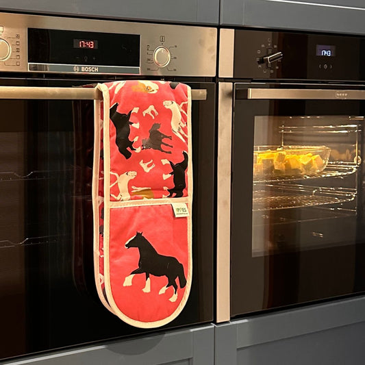 A oink oven mitt with a horse hanging from the handle of built-in oven, with another oven showing food inside in the background.