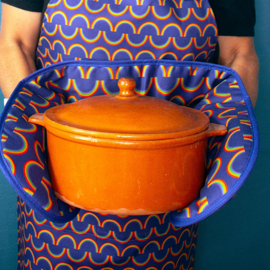 Model wears rainbow stripe apron and holds a hot casserole dish using rainbow stripe oven gloves