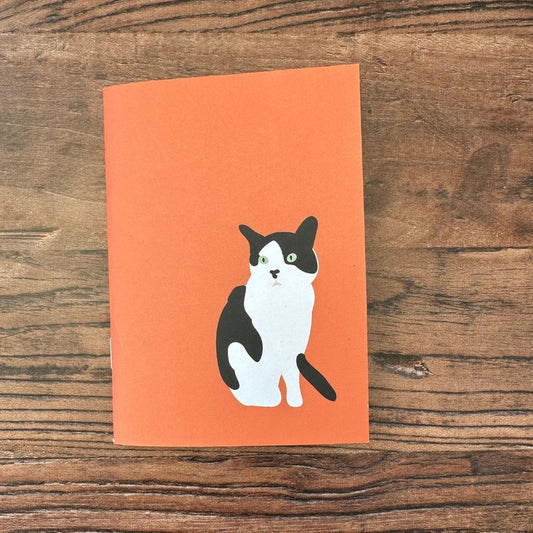 Notebook with a black and white cat illustration on an orange cover, placed on a wooden surface.