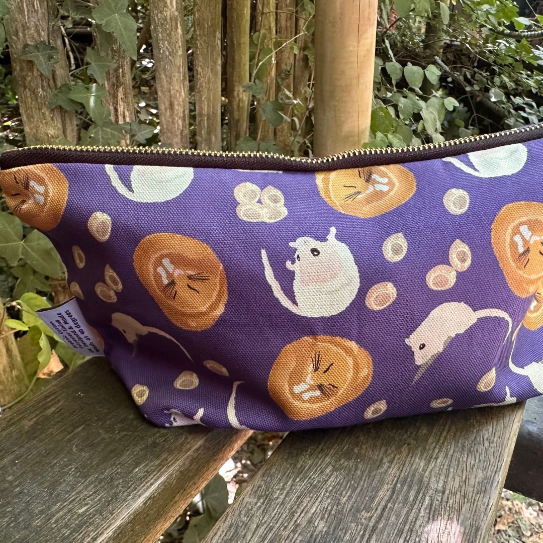 A purple cotton zippered pouch with a playful print of hazel dormice, resting on a weathered wooden bench with a garden fence background.