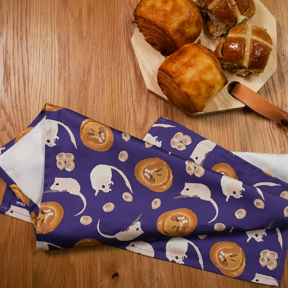 Hot cross buns on a wooden table with a purple tea towel featuring illustrations of hazel dormouse 
