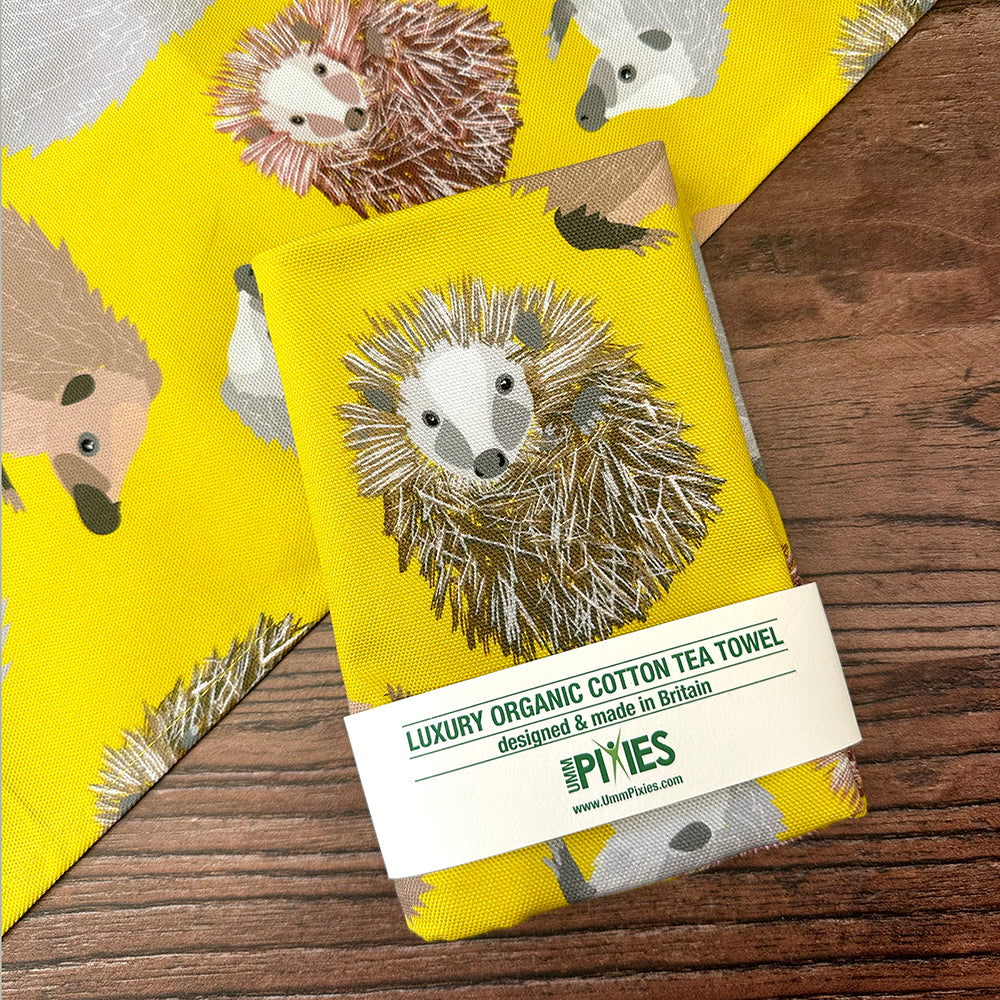 Yellow tea towel with hedgehog design and  UmmPixies branding on a wooden surface.