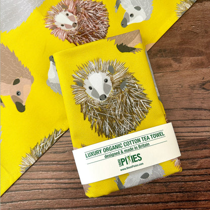 Yellow tea towel with hedgehog design and  UmmPixies branding on a wooden surface.