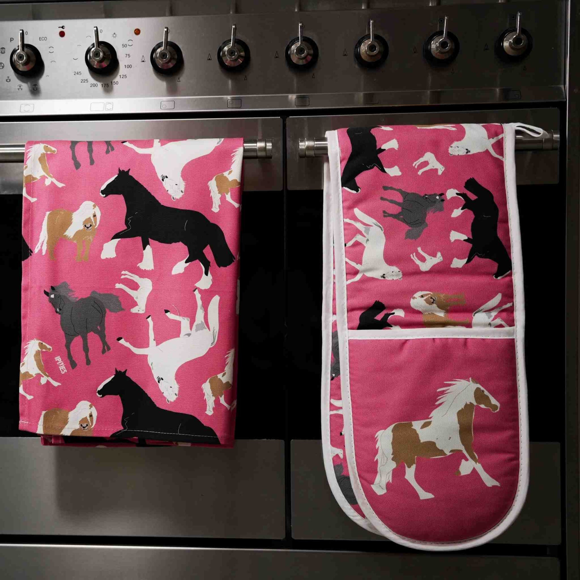 Pink oven gloves and tea towel with horse pattern shown hanging over the handle of a staninless steel stove.