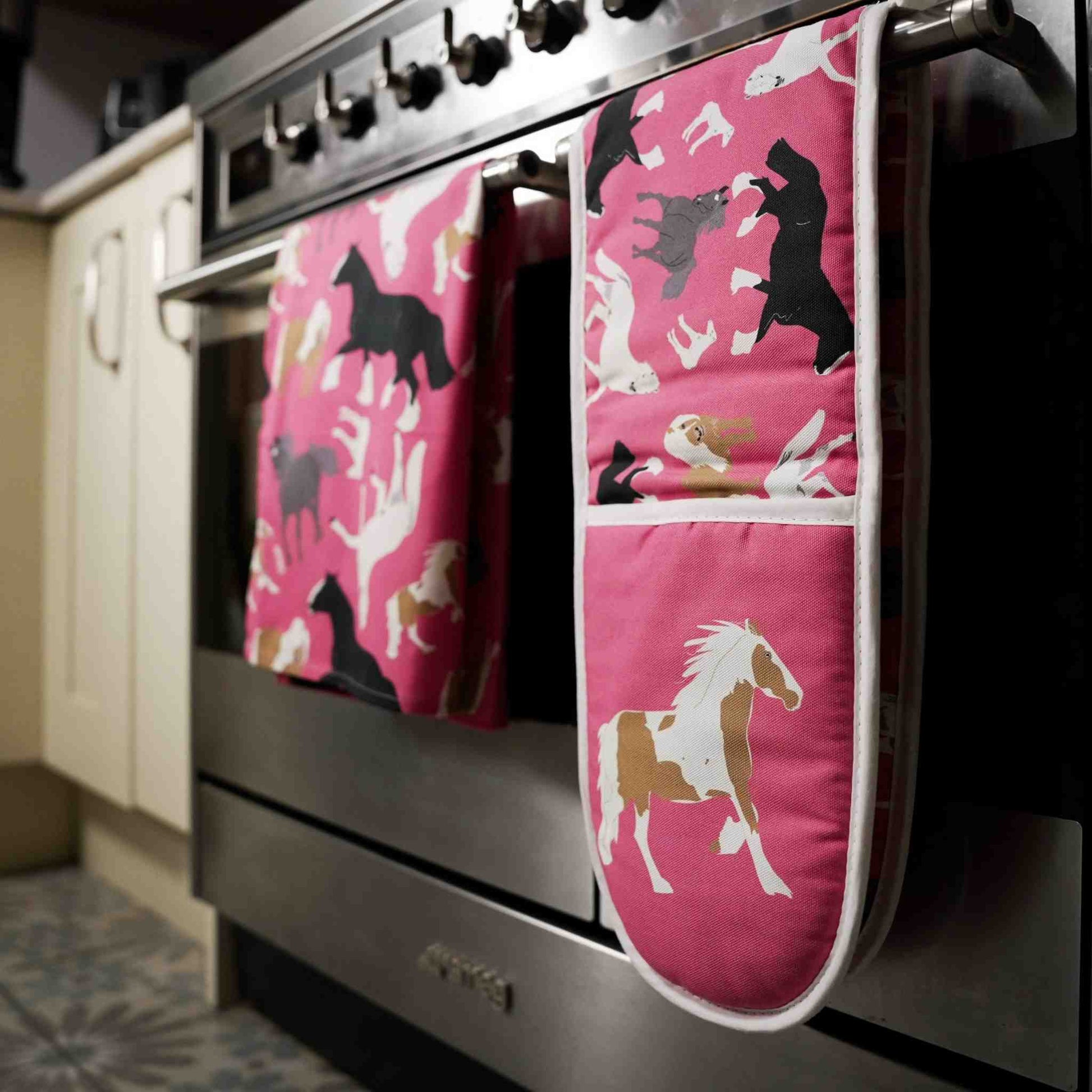 Pink oven mitt and tea towelwith horse designs hanging on a stove.