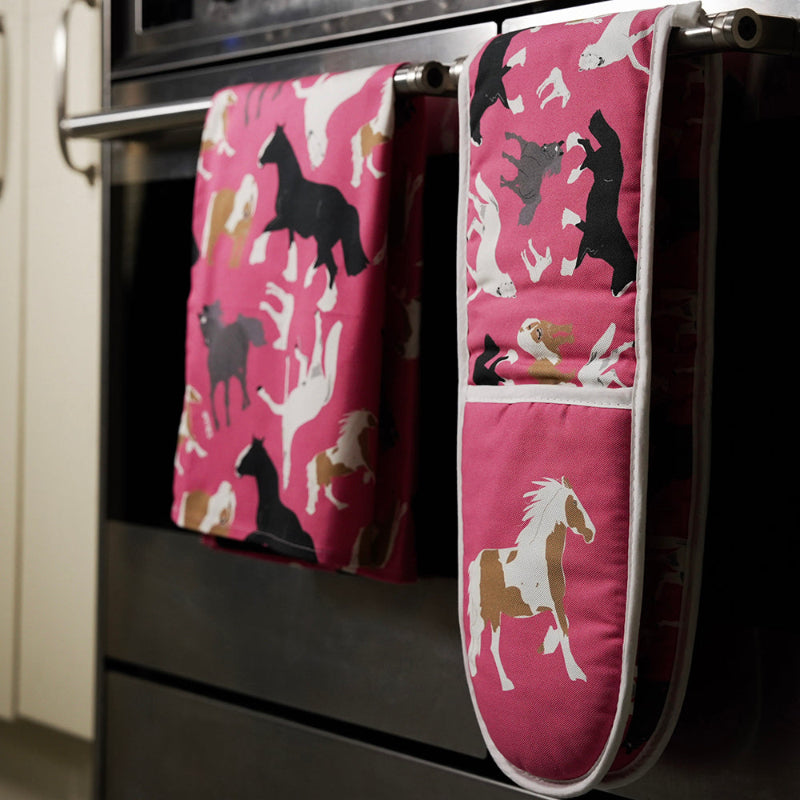 Pink tea towel and matching oven mitts with horse pattern hanging on a stove handle.