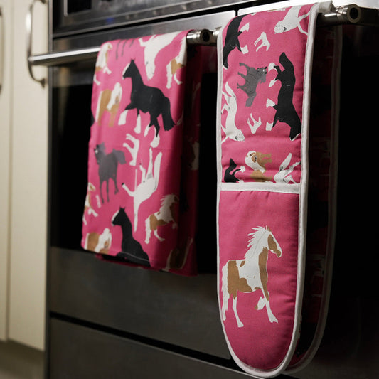 Pink tea towel and matching oven mitts with horse pattern hanging on a stove handle.