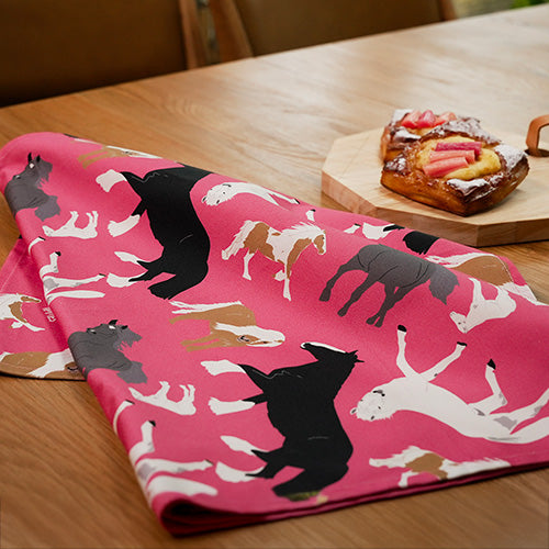 Bright pink horses design tea towel shown on a table next to a board of fruit pastries 