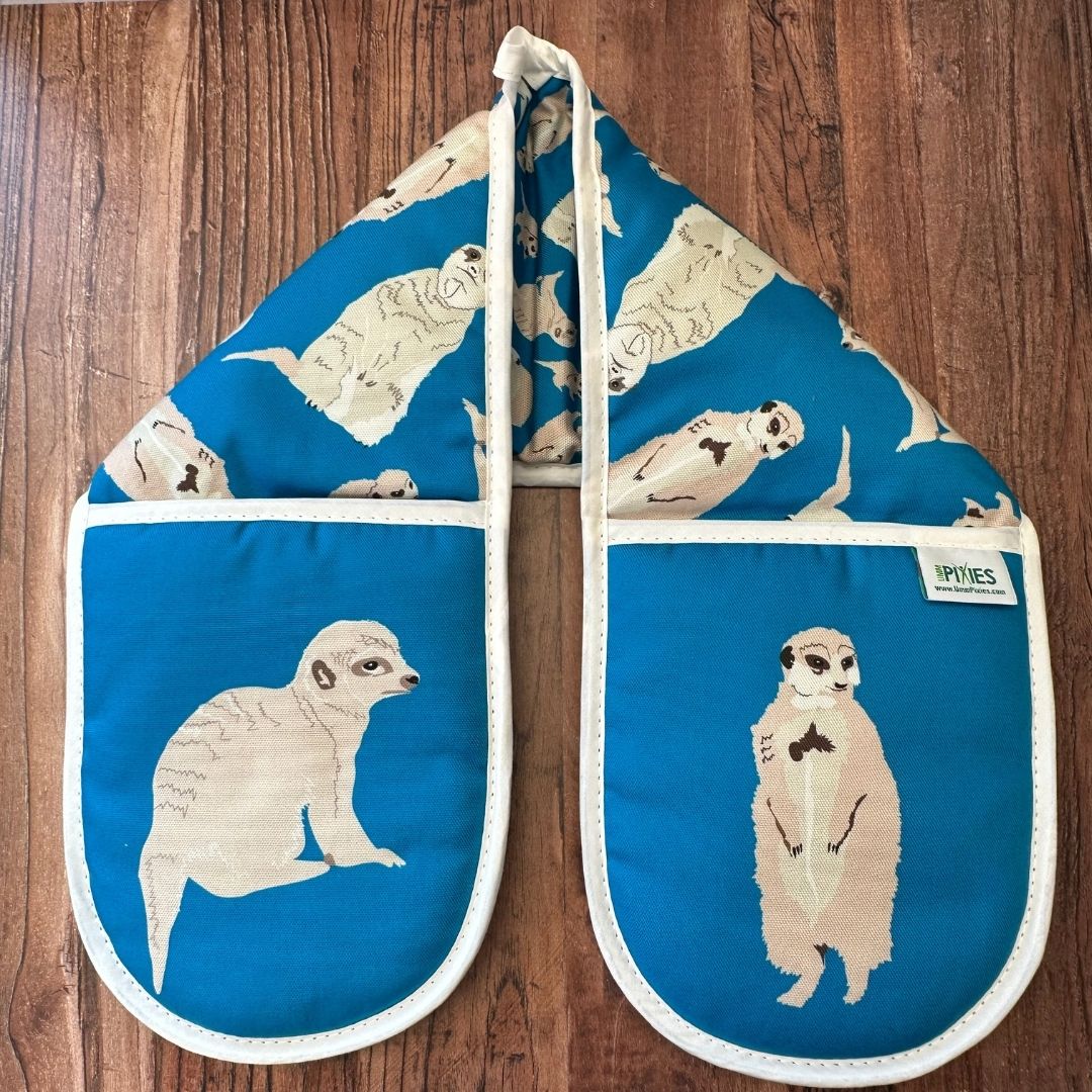 Bright blue cotton oven gloves with meerkat illustrations on a wooden surface