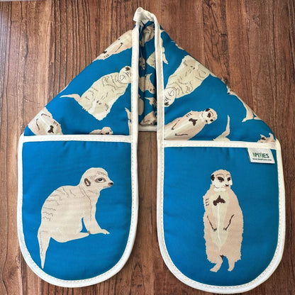 Bright blue cotton oven gloves with meerkat illustrations on a wooden surface