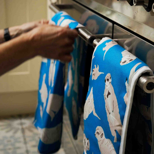 Person hanging a blue tea towel with meerkat design on a stove handle next to a pair of matching oven mitts
