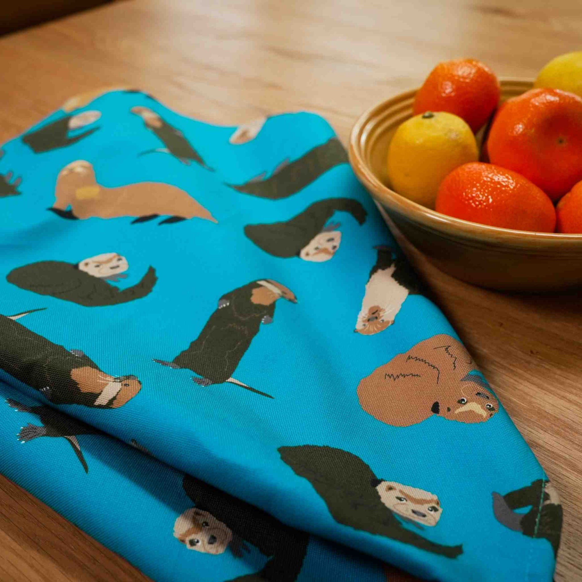 Blue tea towel with otter pattern next to a bowl of fruit on a wooden table