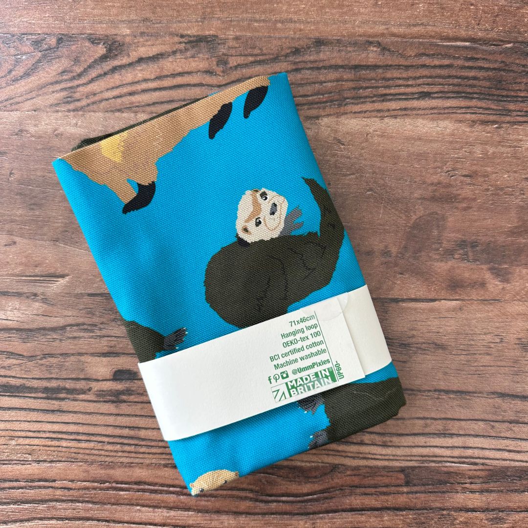 Petrol blue otters design organic cotton tea towel shown in reverse of packaging on wooden table