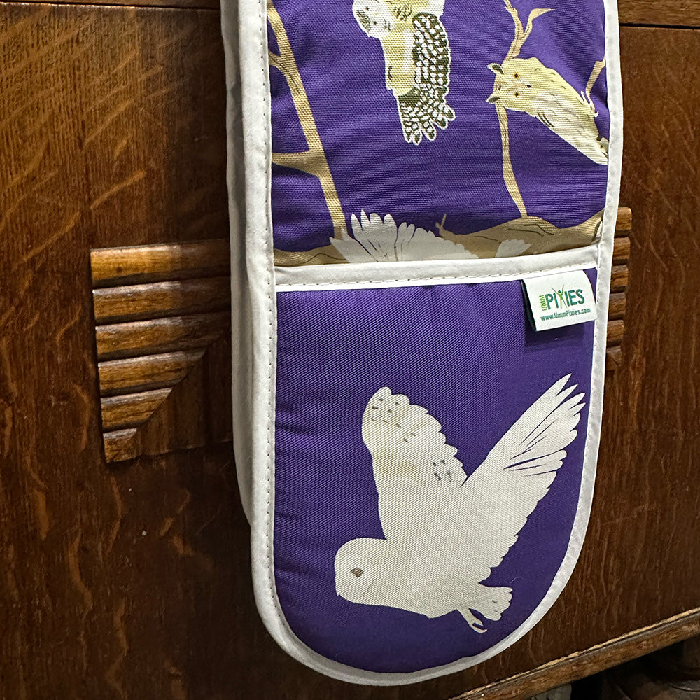 Oven mitt with owl design on a purple background hanging on a wooden cupboard door.