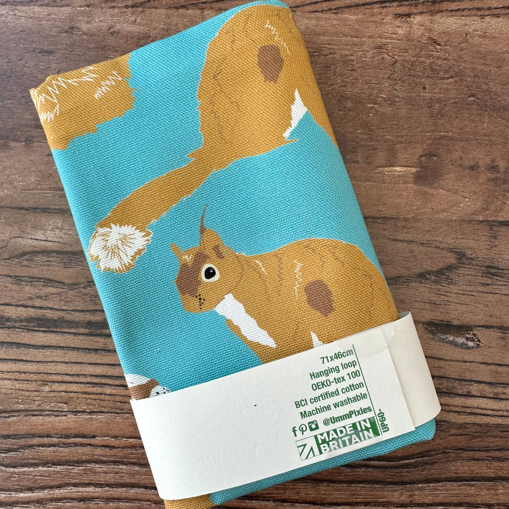Teal cotton tea towel with red squirrel design shown in packaging on a wooden countertop