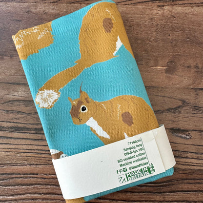 Teal cotton tea towel with red squirrel design shown in packaging on a wooden countertop