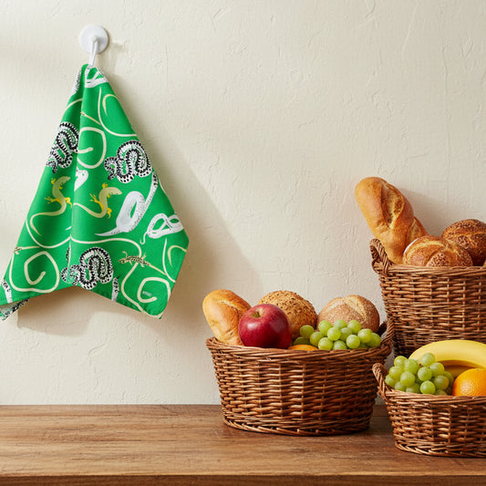 Bright green kitchen tea towel with illustrations of snakes and lizards shown hanging over a counter with baskets of fruit and bread 