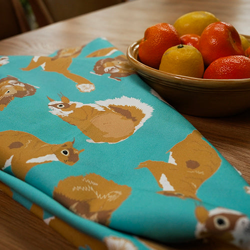 Teal tea towel with squirrel pattern on a wooden table with a bowl of oranges and lemons.