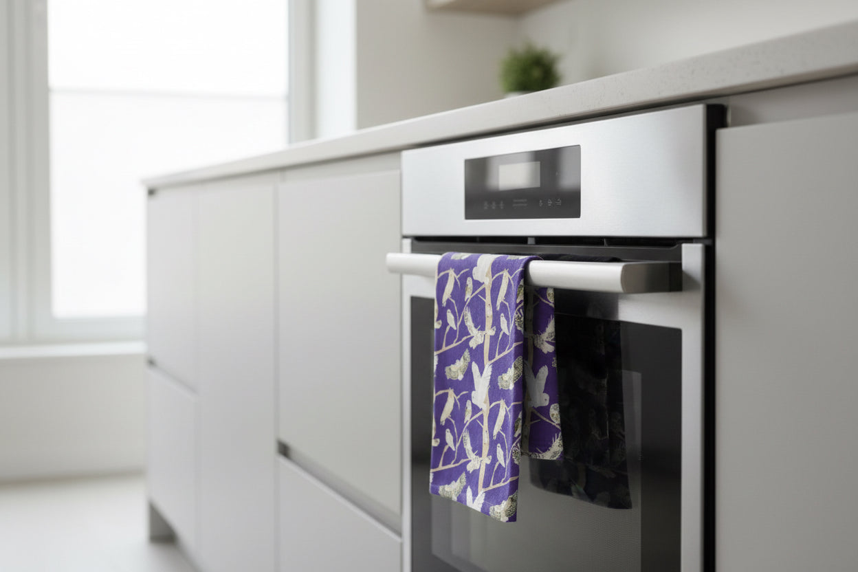 Colourful purple organic cotton tea towel with illustrations of British owls including the bran owl  shown hanging over the door of an oven in a modern kitchen