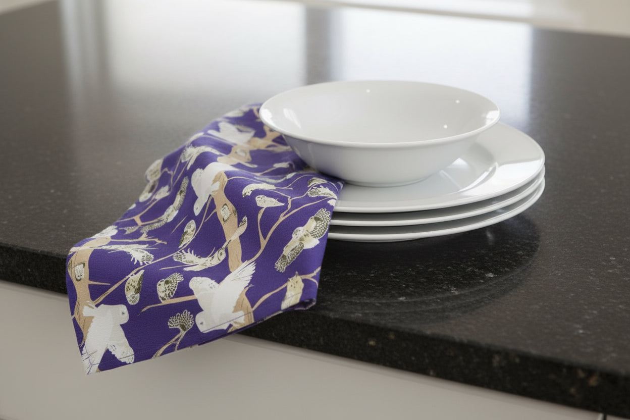 Vibrant purple cotton tea towel featuring illustrations of native British Owls shown with a stack of clean dishes 