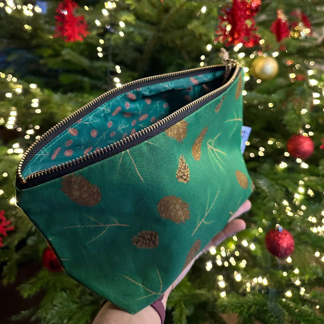 Green fabric zipped pouch with evergreen cones and sprigs illustrations patterns held in front of a decorated Christmas tree.