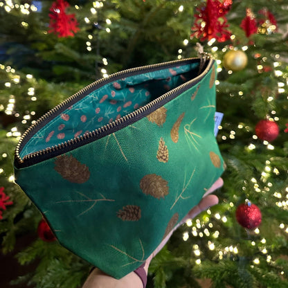 Green fabric zipped pouch with evergreen cones and sprigs illustrations patterns held in front of a decorated Christmas tree.