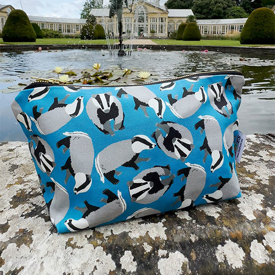 Blue waterproof lined zip pouch with badger pattern on a stone ledge by a pond with a building in the background