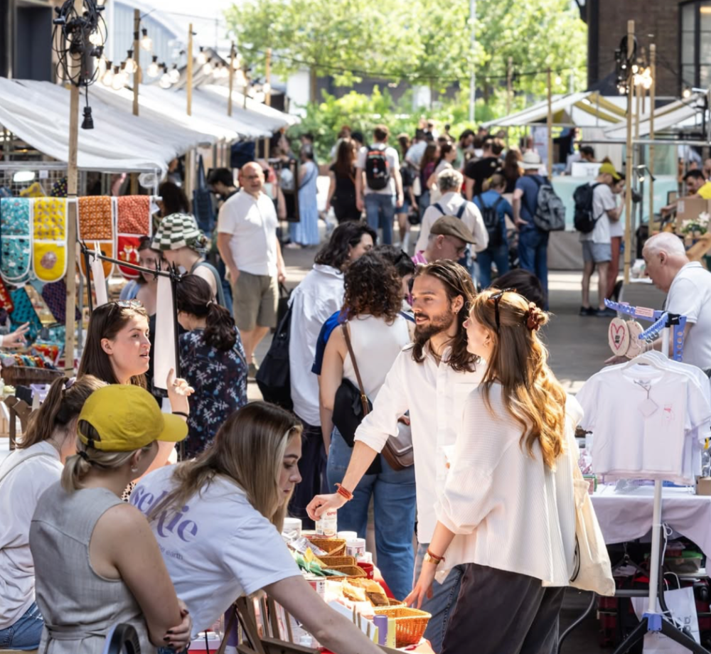 People shopping at an outdoor market with stalls and people walking around.UmmPixies oven gloves display visible on left side