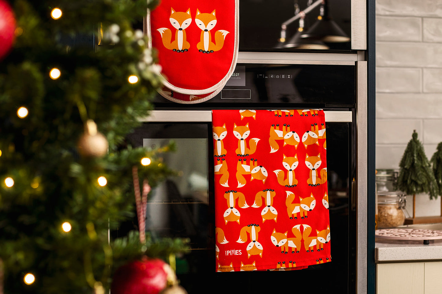 Red kitchen towel and macthing oven gloves with fox pattern hanging on a kitchen oven door next to a Christmas tree.