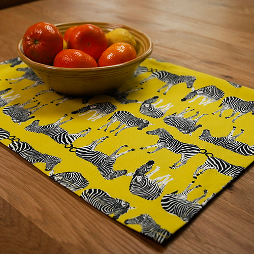 Yellow tea towel with zebra pattern used as a placemat for a bowl of oranges on a wooden table.