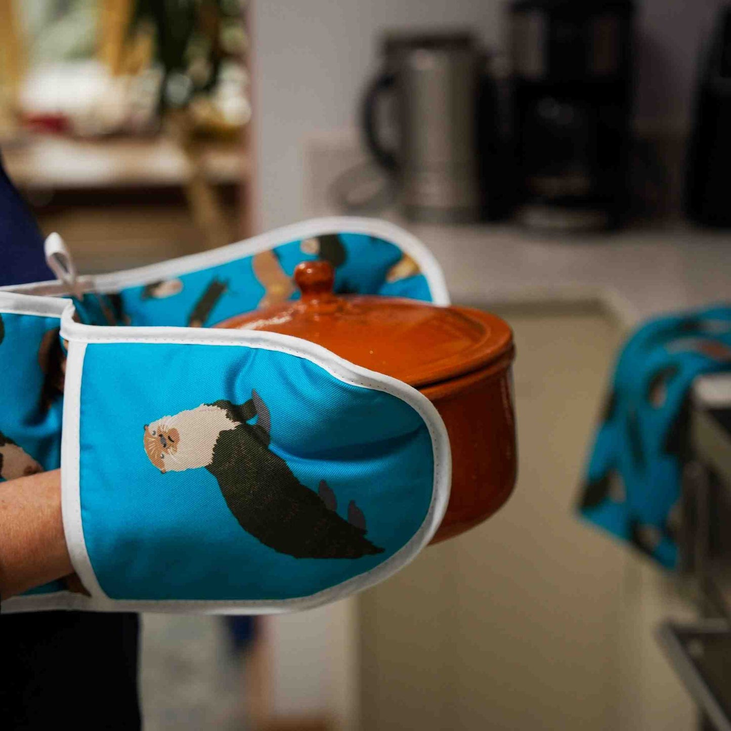 Person holding a pot with a blue oven mitt featuring an otter design in a kitchen.