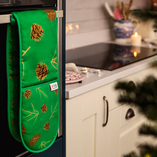 A pair of green double oven mitt with illustrations of Douglas fir cones and spruce sprigs shown handing over an oven door handle